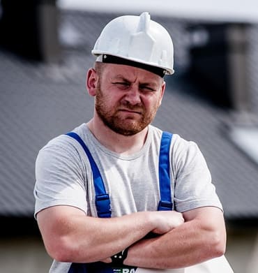 Construction worker wearing a white hard hat and safety harness, standing outdoors.